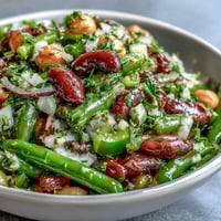A vibrant Three-Bean Salad with green beans, kidney beans, and chickpeas tossed in a tangy apple cider vinaigrette, served chilled in a clear glass bowl.  